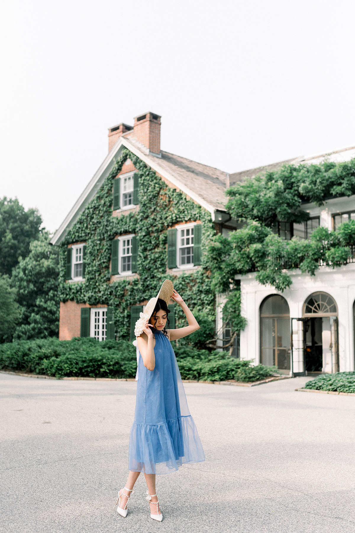 In her Anthropologie French blue gown and a hat adorned with delicate white hydrangeas, Karla adds a touch of vintage charm to the scene as she poses gracefully in front of a historical house at Longwood Gardens.