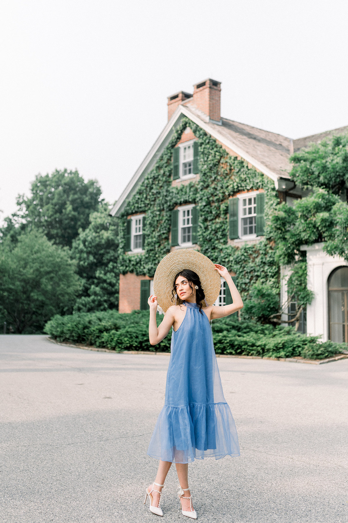 Bride exudes timeless elegance as she poses in front of a historic house at Longwood Gardens, dressed in the Anthropologie French blue gown and a charming white hydrangea-adorned hat.