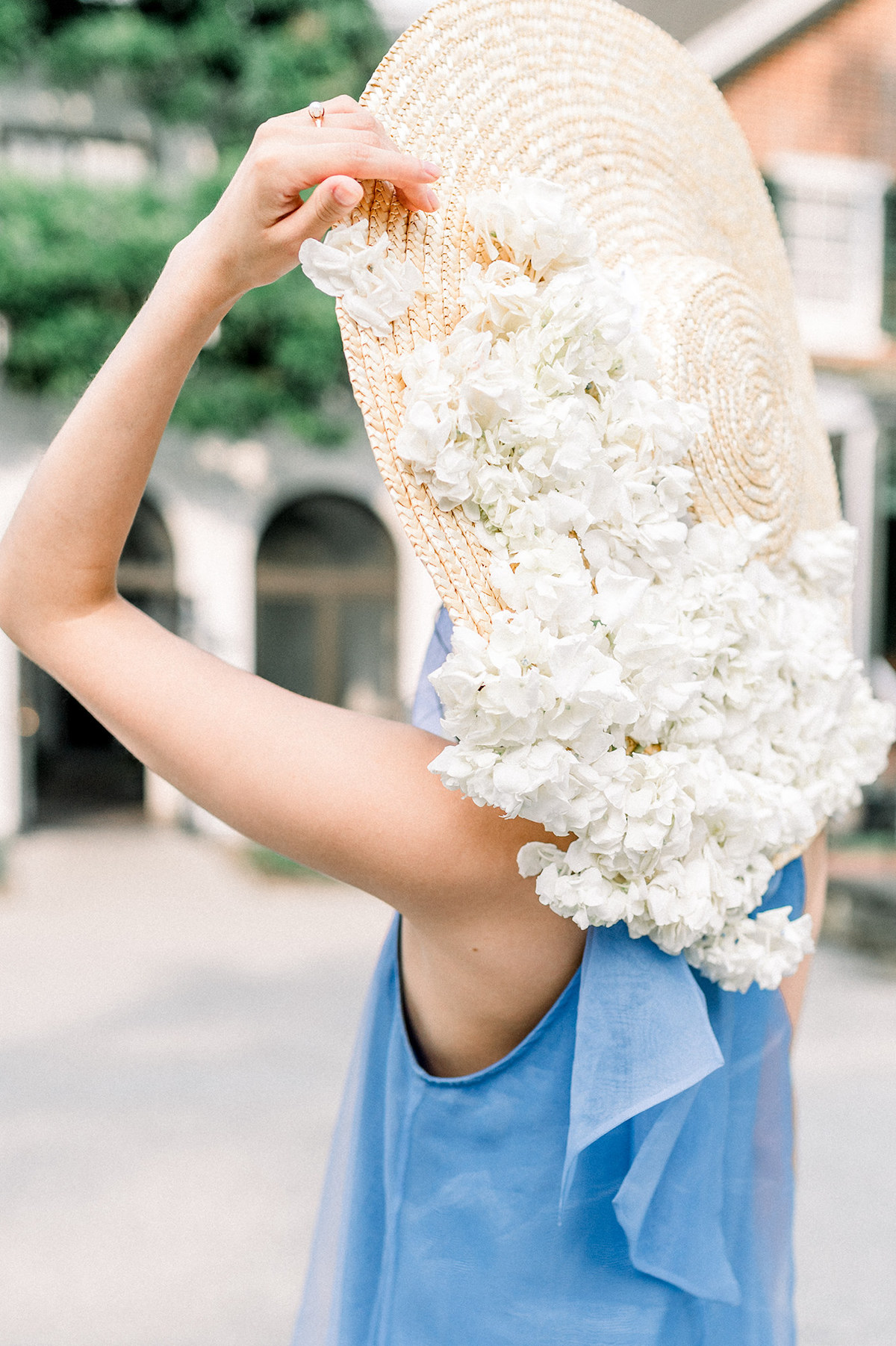 A picturesque moment captured as Bride, donning the Anthropologie French blue gown and a boat-style hat with white hydrangeas, strikes a pose against the backdrop of a historic house at Longwood Gardens.