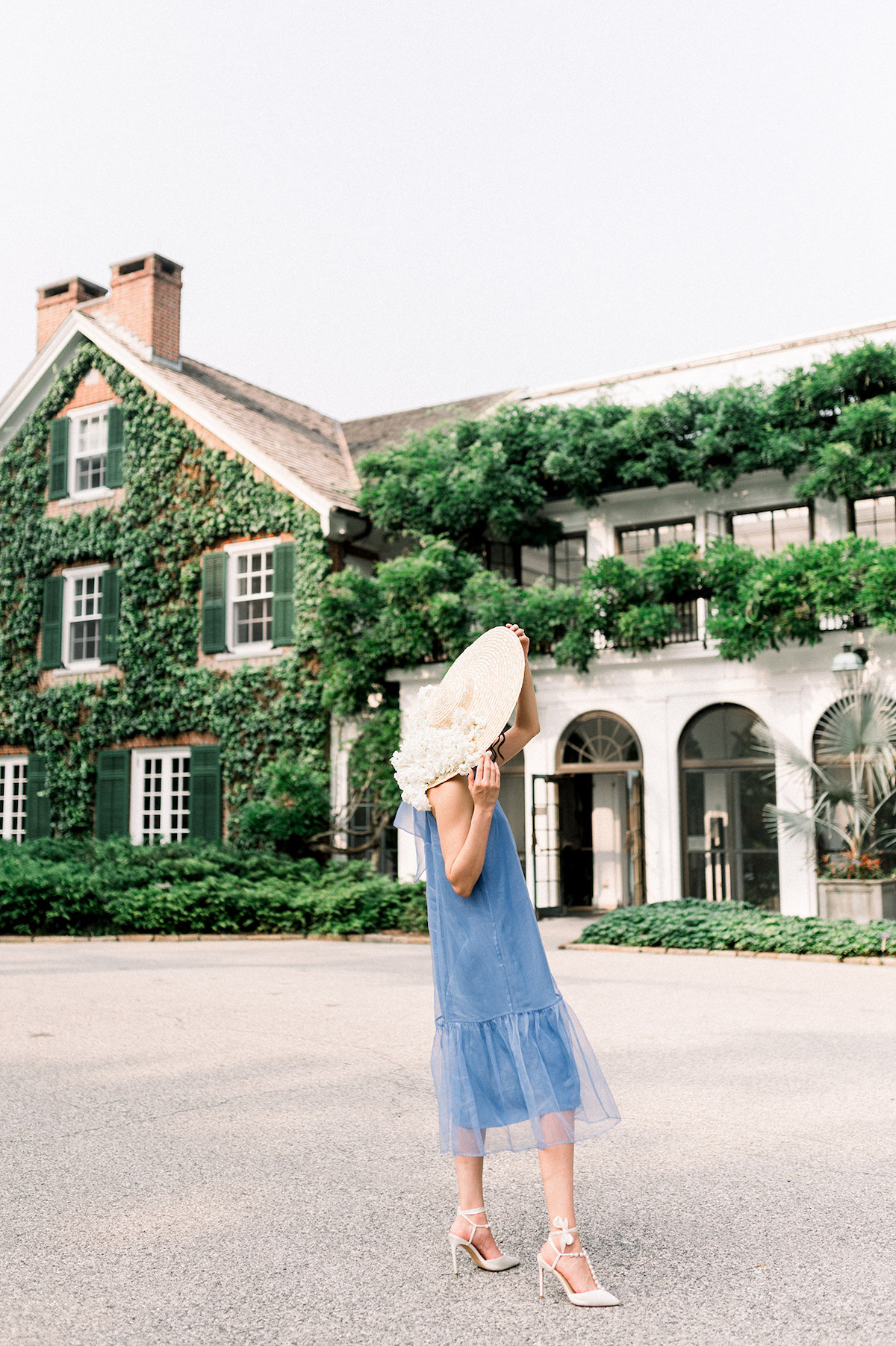 In her Anthropologie French blue gown and a hat adorned with delicate white hydrangeas, Karla adds a touch of vintage charm to the scene as she poses gracefully in front of a historical house at Longwood Gardens.
