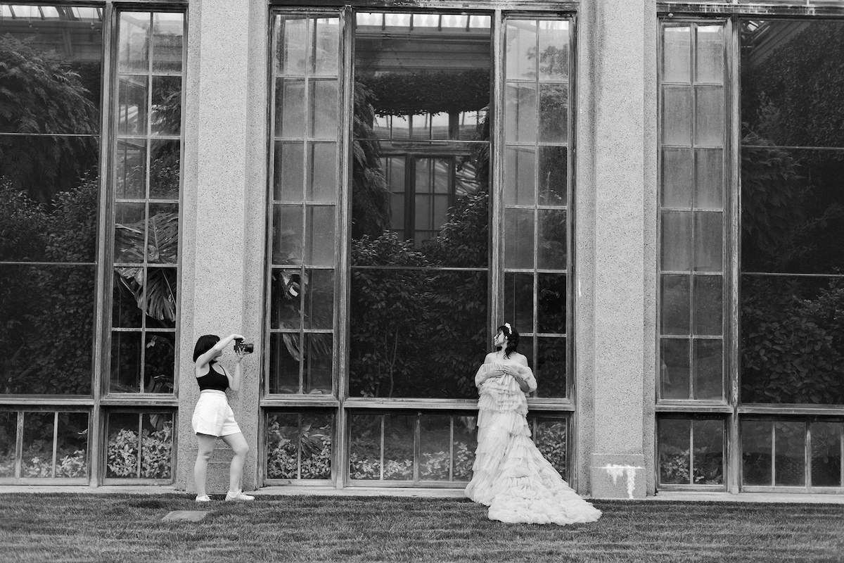 A candid behind-the-scenes shot of April Raymond at work, skillfully photographing Karla in her mauve couture gown in the garden.